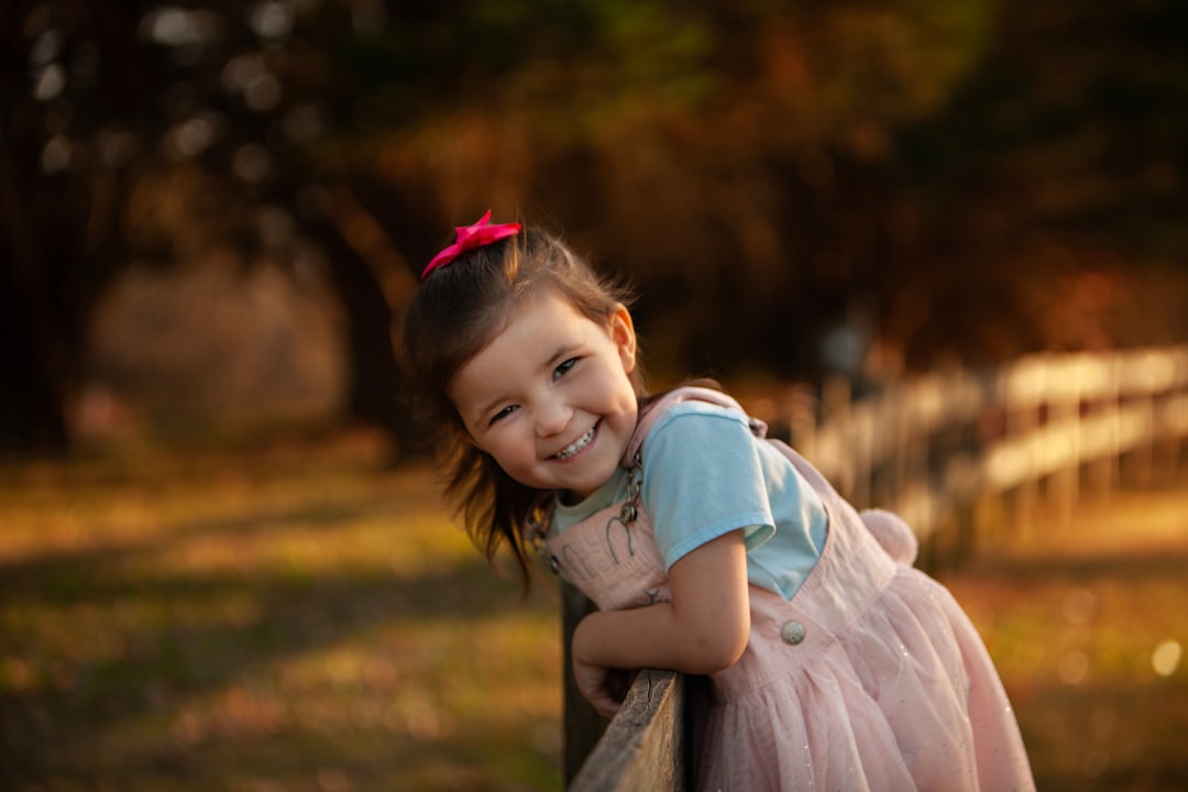 A happy young girl leans on a wooden fence outdoors.